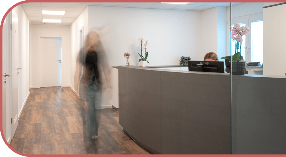 A busy office or medical reception area featuring a large curved desk, a seated receptionist, and a blurred figure walking down a nearby hallway.